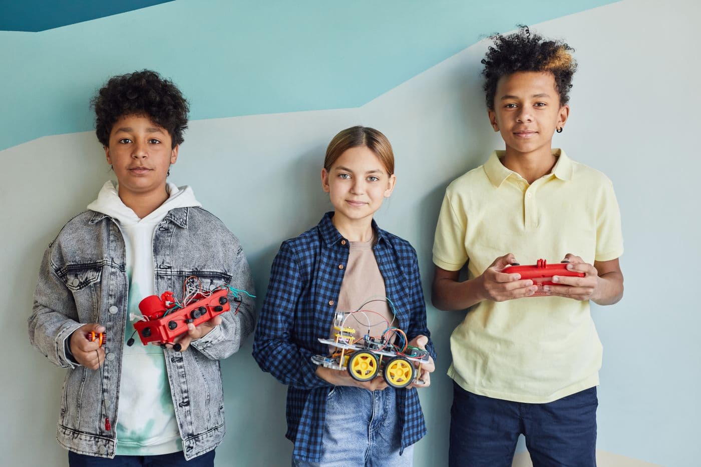 Three students standing against a blue wall, each holding a robot they built.